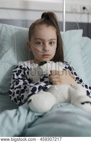Sick Kid Wearing Oxygen Tube While Sitting In Patient Bed Having Plush Bear Toy. Under Treatment Ill