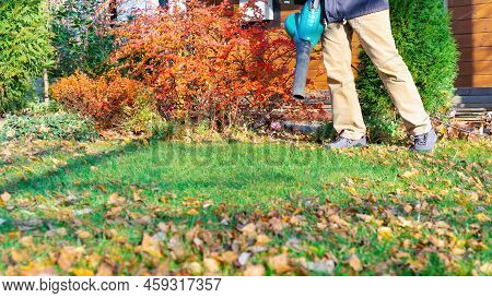 A Gardener With A Blower Cleans The Lawn From Fallen Leaves Against The Backdrop Of A Beautiful Land