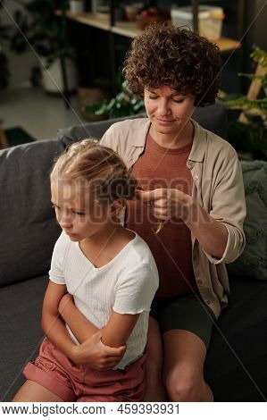 Young Mother In Casualwear Plaiting Hair Of Her Cute Youthful Daughter