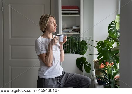 Tranquil Middle-aged Female Enjoys Hot Drink From Mug Standing At Windowsill. Mature Blonde Woman En