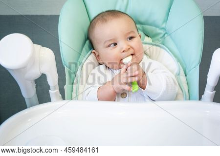 A Hungry Baby Is Gnawing On A Plastic Spoon At The Table On A High Chair. Teething, Whims, Itchy Gum