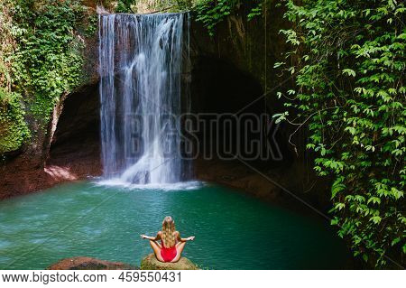 Woman In Bikini Sit On Rock, Meditating In Yoga Pose Under Falling Water Of Suwat Waterfall In Tropi