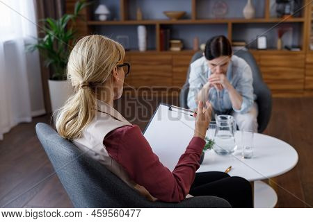 Over Shoulder View Of Female Psychologist Sitting In Armchair, Talking With Upset Woman Patient. Psy