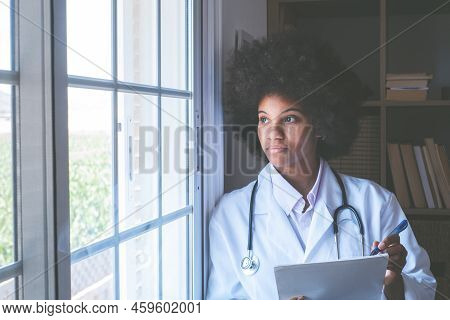 Thoughtful Female African American Doctor With Stethoscope, Documents And Pen Looking Outside Her Cl