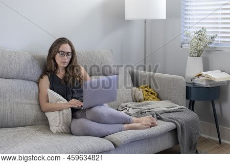 Mid Age Woman With Long Hair Sitting On The Couch With Laptop Computer In The Gray Modern Room. Work