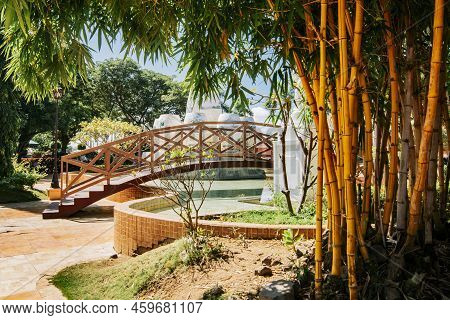 Wooden Bridge In A Park Over Water Fountain Surrounded By Bamboo. Side View Of A Small Wooden Bridge