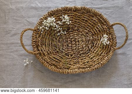 Top View Of Brown Woven Basket With Two Handles And Tiny White Flowers On Linen Fabric Background