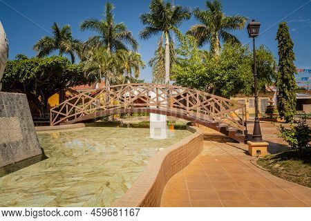Side View Of A Small Wooden Bridge Over A Water Fountain In A Calm Park. Nagarote Central Park.  Vie