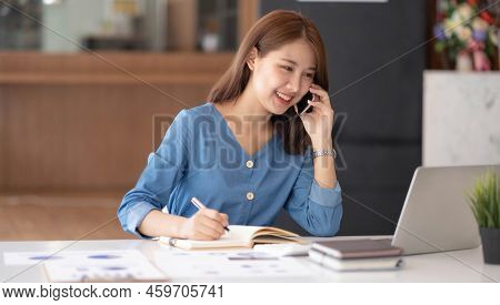 Smiling Pretty Girl Working On Laptop Computer While Sitting At Office Table, Taking Notes In A Diar