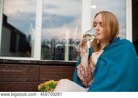 Portrait Of Young Buxom Woman Covered By Blue Blanket Sitting Near Cut Fruits On Tray, Drinking Glas