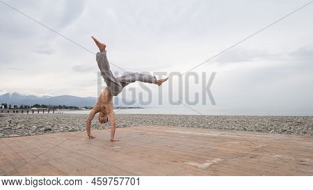Shirtless Caucasian Man Doing Backflip On Pebble Beach.