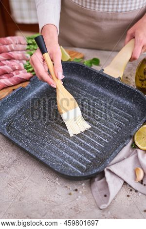 Woman Smearing Oil On A Grill Pan To Cook Aspargus Covered With Bacon