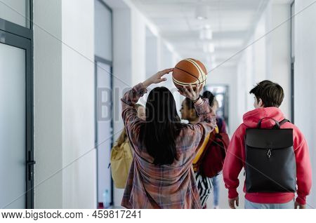 Rear View Of Young High School Students Walking In Corridor At School With Basketball Ball, Back To 