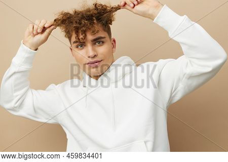 A Stylish Young Man In A White Hoodie Stands On A Beige Background And Pulls His Curly Hair Aside Wi