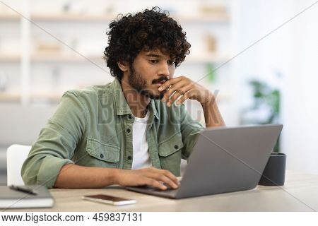 Pensive Dark-skinned Guy Sitting At Workdesk In Front Of Laptop