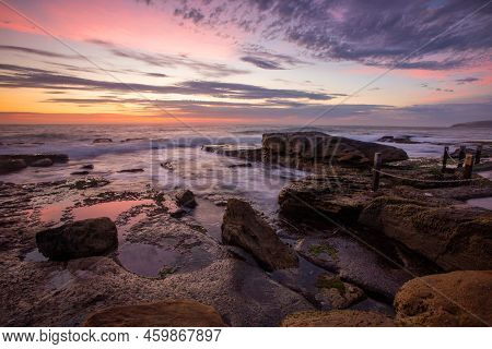 A Pretty And Colourful Sunrise Is Reflected On The Rocks And Waves Cascaed And Flow Across The Rocky