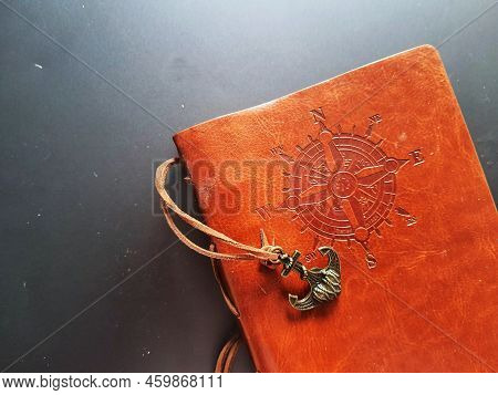 A Notebook With A Brown Leather Cover With A Compass Motif And Anchor Strap.