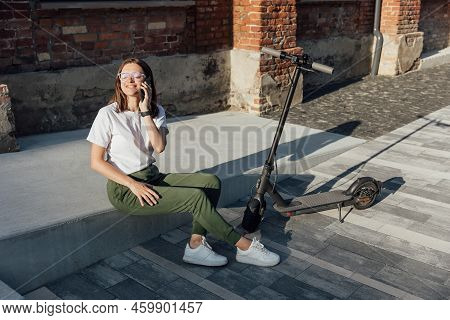 Young Female Sitting On Urban Stairs And Speaking On Smartphone Near Electric Scooter