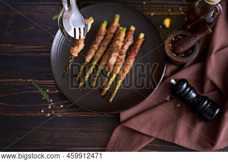 Baked Aspargus Covered With Bacon On A Black Ceramic Plate On Wooden Kitchen Table