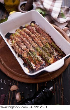 Ceramic Baking Dish With Baked Aspargus Covered With Bacon On Wooden Kitchen Table