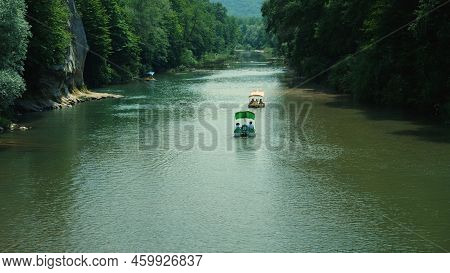 Pedal-powered Catamarans Float Down The River. Dense Green Trees Grow Along The River Banks