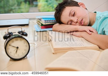 Blurred Black Alarm Clock On The Background Of A Tired Sleepy Schoolboy Falling Asleep On A Textbook