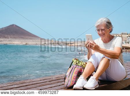 Happy Senior Woman Sitting Close To The Beach While Using Mobile Phone  - Elderly Beautiful Lady Enj