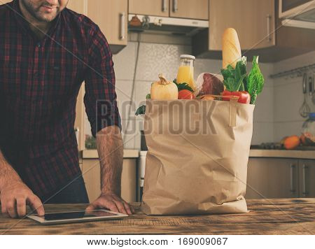 Full paper bag of different vegetables and fruit on a wooden table close up. In the background a man is shopping via the Internet using the tablet