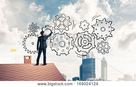 Young businessman standing on house roof and drawing gear mechanism. Mixed media