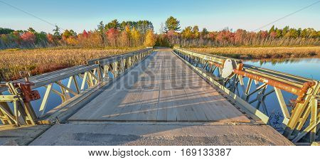 Wide angle view of clear blue sky, late afternoon sunshine, casts long shadows on a light steel and timber bridge, single point perspective over a lake in Ontario, Canada.