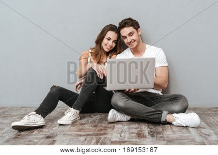 Couple sitting on the floor with laptop computer in studio. Full length image. Isolated gray background