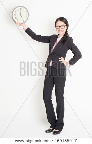 Full length front view of young Asian businesswoman in formalwear hand holding clock at 5pm, standing on plain background.
