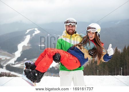Photo of happy loving couple snowboarders on the slopes frosty winter day. Look at camera. Man holding lady on hands.