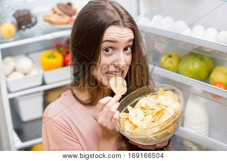 Young woman eating potato chips near the refrigerator full of healthy food