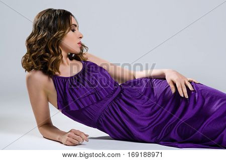 Beautiful brunette in purple dress lying on light background
