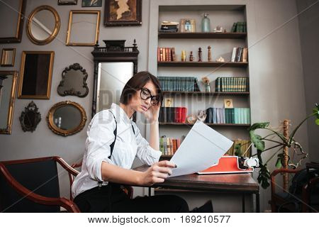 Side view of attractive authoress in glasses and white shirt sitting by the table with paper and typewriter. Bookshelf on background