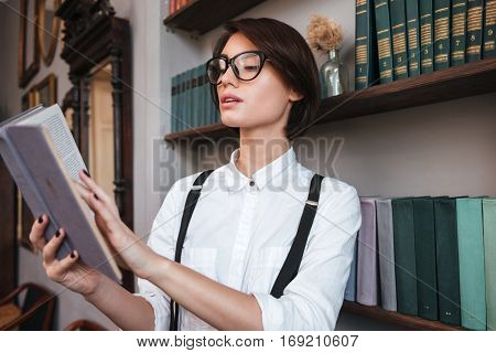 Authoress in glasses and shirt reading book near the bookshelf