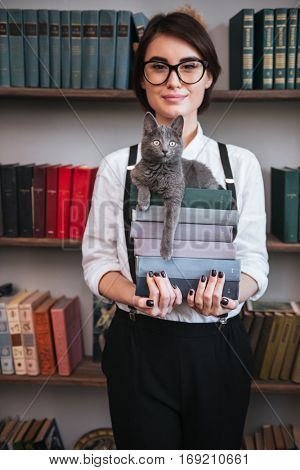 Vertical image of Smiling Authoress in glasses and white shirt holding books with cat on them and looking at camera