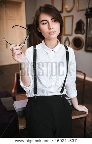 Vertical image of Authoress in shirt standing near the table and holding eyeglasses in hand