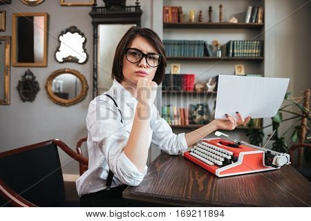 Side view of pensive authoress in glasses and white shirt sitting by the table with paper and typewriter and looking at camera
