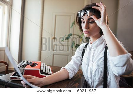 Side view of beauty authoress in white shirt holding her eyeglasses and sitting by the table with paper and typewriter