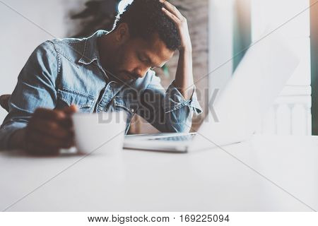 Tired young African man using laptop while sitting at the table on a sunny morning.Concept of people working hard home.Blurred background, flare effect