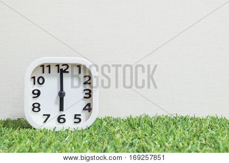 Closeup white clock for decorate in 6 o'clock on green artificial grass floor and cream wallpaper textured background with copy space