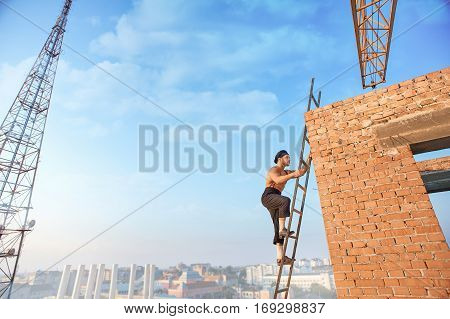 Side view of handsome builder with bare torso in hat climb up ladder up. Ladder leaning on brick wall at un finished building. High TV tower and cityscape on background.