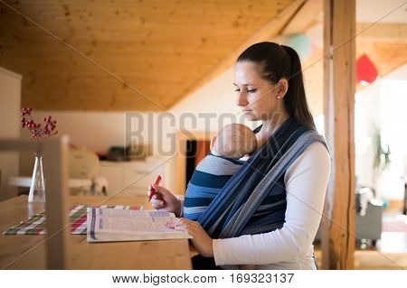 Beautiful young mother at home with her baby son sleeping in sling, reading a book or studying something