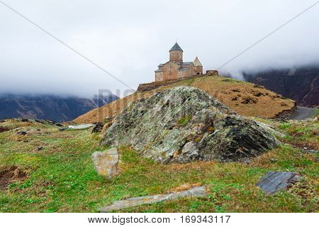 Gergeti Trinity Church Or Tsminda Sameba in the morning mist near village Stepantsminda, Georgia. Mount Kazbegi. Spring Or Summer Season.