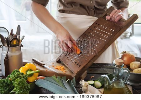 Woman grate carrots on vintage grater on wooden table near window. Rustic style
