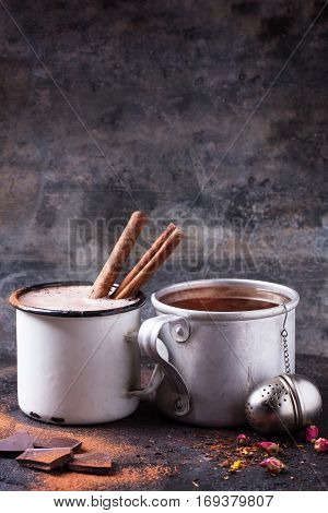 Two vintage mugs with tea and hot chocolate, served with dry tea, rose buds, chopped chocolate and cinnamon sticks over black background
