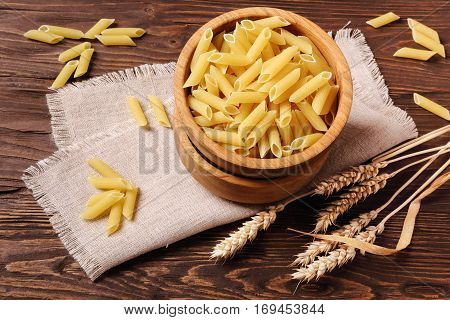 Raw pasta penne and rye ears in a wooden bowl on the table