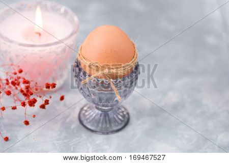 Easter brown egg in a crystal blueish cup, burning pink candle in the background,tender red psring flowers,blue and white concrete background,decoration concept